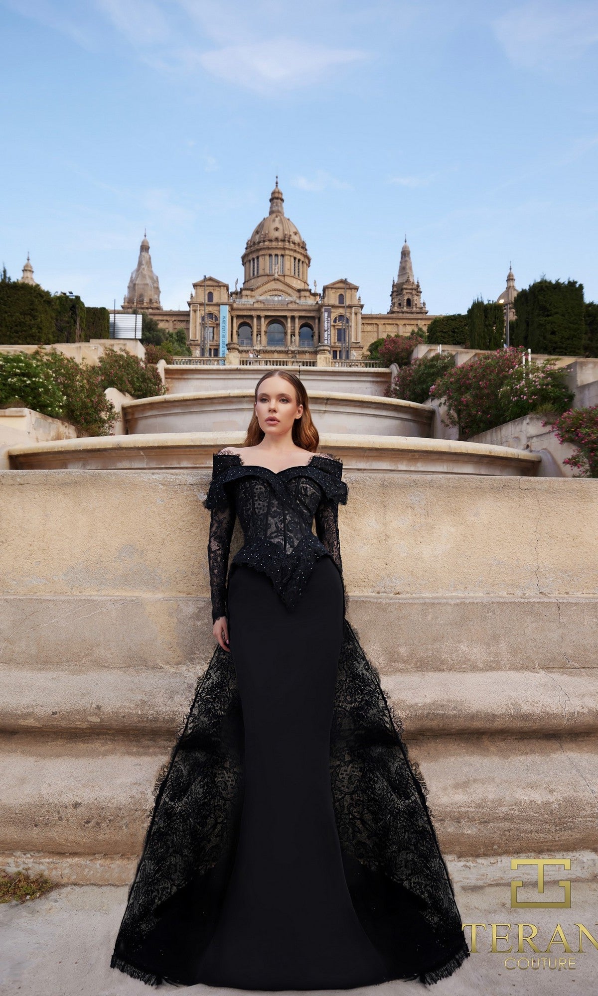 A woman in a black Long Formal Dress (Terani 261E6208) stands on stone steps before an ornate historic building under a blue sky.